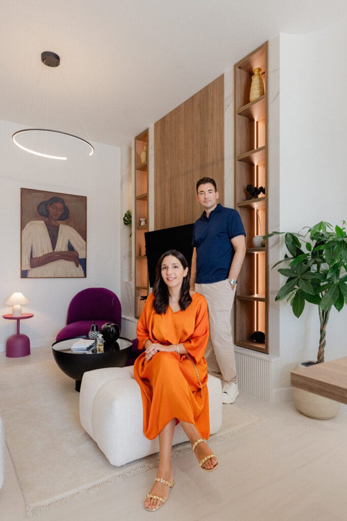 Woman in an orange dress seated on a white lounge with a man standing behind in a modern living room with built-in wood shelves and plants.